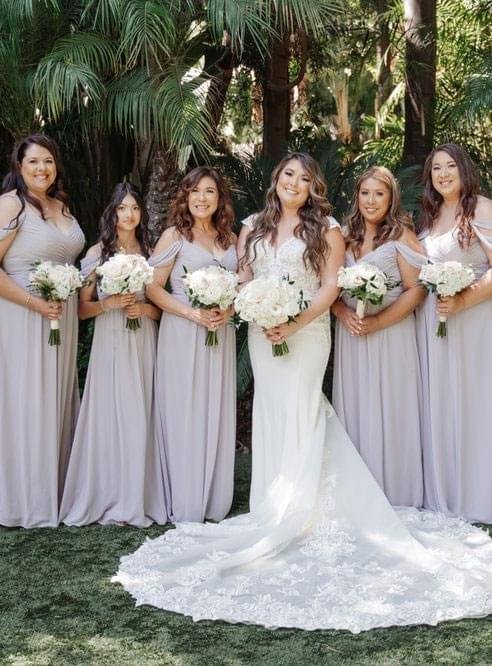 A bride in a white gown and her bridesmaids in matching gowns pose together in front of lush palm trees.